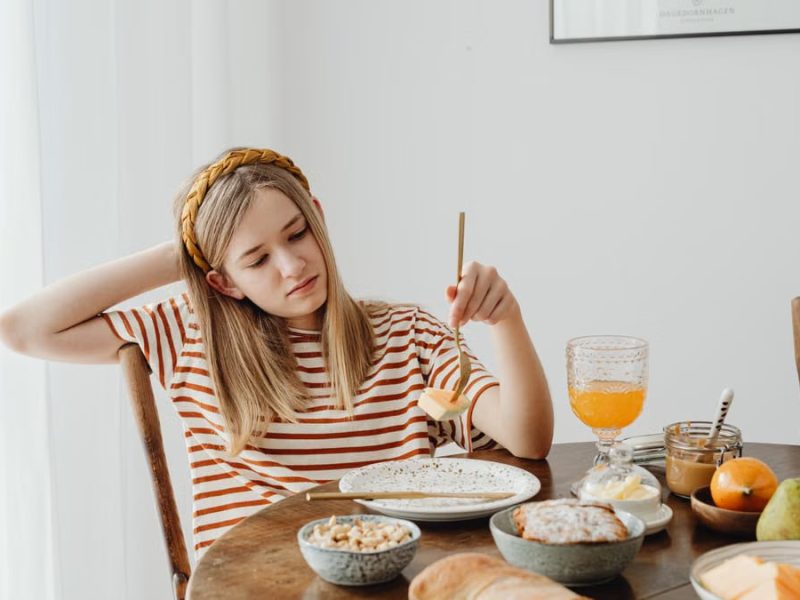 Teen girl in striped shirt at breakfast table with fruits, bread, and juice. Casual morning setting.