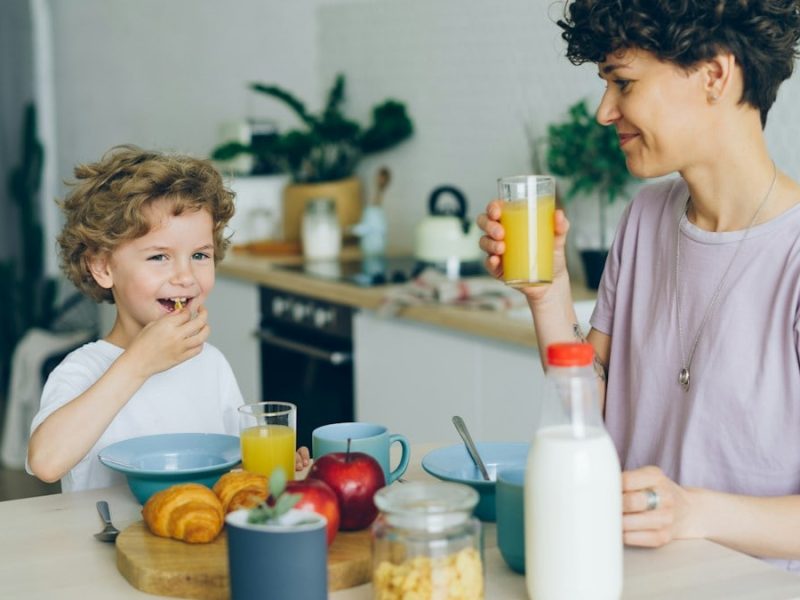 Young woman drinking orange juice while son eating cereal at table in kitchen
